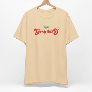 May include: A beige short-sleeved t-shirt featuring the word Groovy printed in red across the chest. The letter o in the word is replaced by a graphic of two red cherries with a green stem and leaves positioned above the text on the shirt.