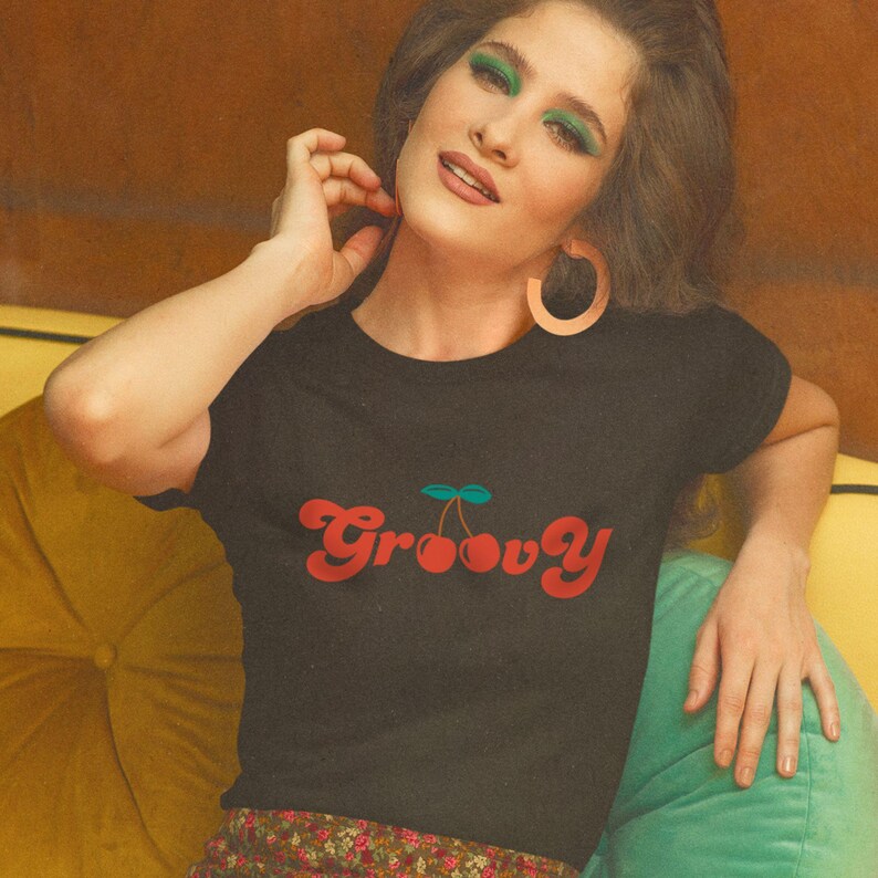 May include: A black short-sleeved t-shirt featuring the word Groovy in a red retro-style font, with two red cherries and a green stem replacing the double o in the text. The shirt has a crew neckline and a simple, fitted silhouette.