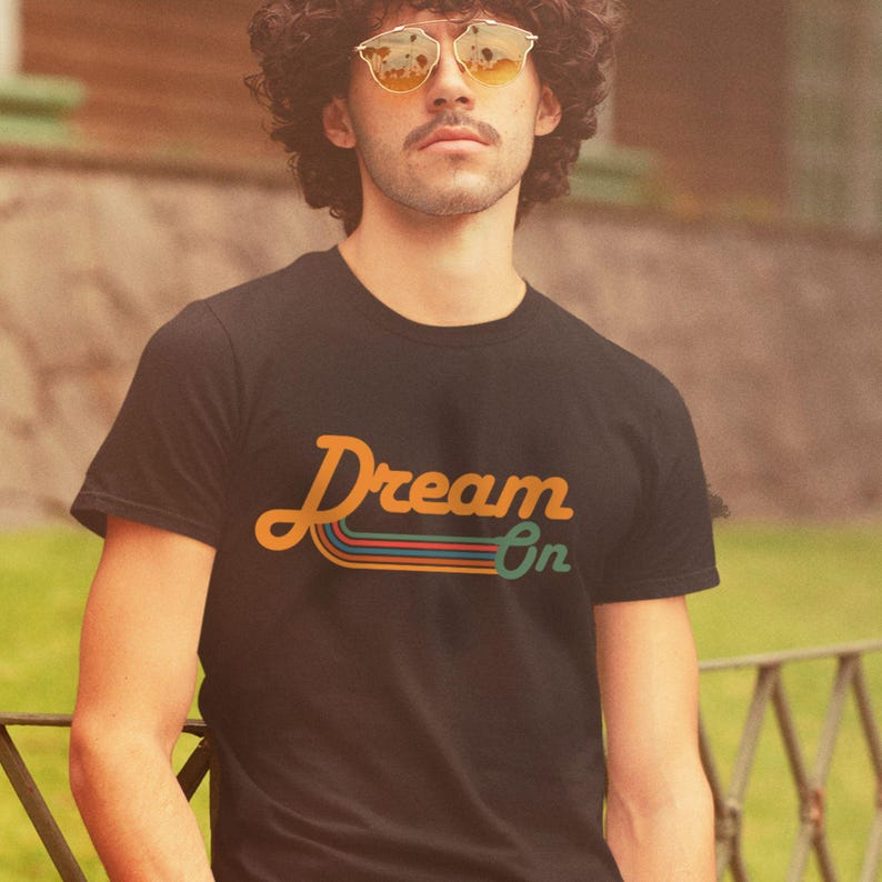 K&ouml;nnte beinhalten: Ein schwarzes T-Shirt mit einer Retro-Grafik, auf der "Dream On" in orangefarbenen und gelben Buchstaben mit bunten Streifen darunter steht.