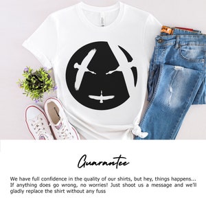 Pu&ograve; includere: Maglietta bianca con un grafico circolare nero che presenta due silhouette di oche in volo. Le oche volano in direzioni opposte, creando un'illusione ottica di un solo uccello. Il testo "Guarantee" &egrave; scritto sotto l'immagine.