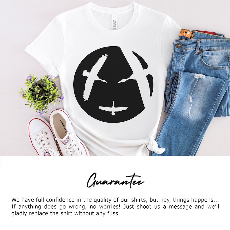 Pu&ograve; includere: Maglietta bianca con un grafico circolare nero che presenta due silhouette di oche in volo. Le oche volano in direzioni opposte, creando un'illusione ottica di un solo uccello. Il testo "Guarantee" &egrave; scritto sotto l'immagine.