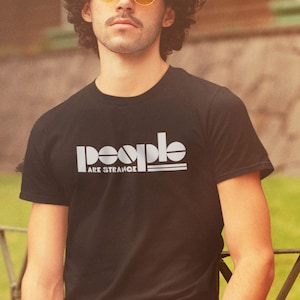 May include: Black crew neck t-shirt with the white text "people ARE STRANGE" in a bold, stylized font. The shirt is a solid color and appears to be made of cotton or a similar material. The design is centered on the chest.