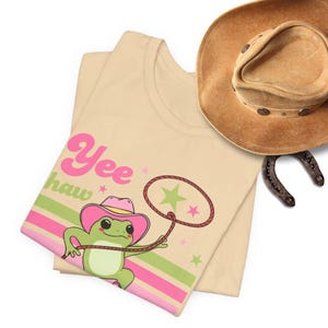 Pu&ograve; includere: Una maglietta beige con una rana di cartone animato che indossa un cappello da cowboy rosa e fa il lazo a una stella. Il testo "Yee haw" &egrave; stampato sulla maglietta in lettere rosa e verdi.