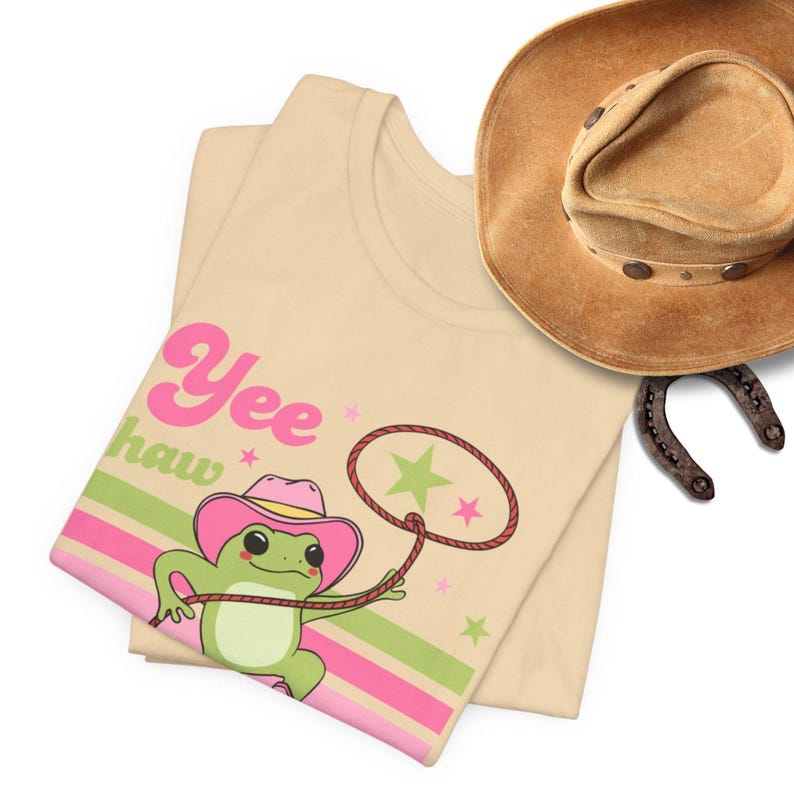 Pu&ograve; includere: Una maglietta beige con una rana di cartone animato che indossa un cappello da cowboy rosa e fa il lazo a una stella. Il testo "Yee haw" &egrave; stampato sulla maglietta in lettere rosa e verdi.