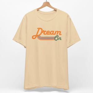 K&ouml;nnte beinhalten: Ein beiges T-Shirt mit einer Retro-Grafik, auf der "Dream On" in Orange mit gr&uuml;nen und orangefarbenen Streifen steht.