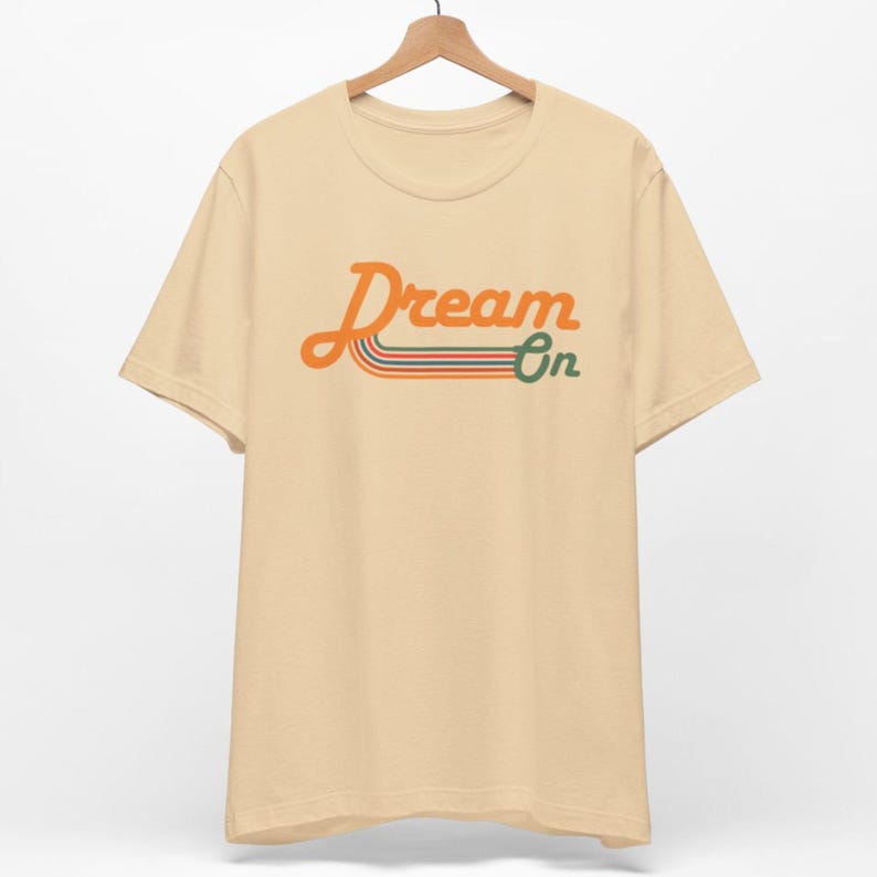 K&ouml;nnte beinhalten: Ein beiges T-Shirt mit einer Retro-Grafik, auf der "Dream On" in Orange mit gr&uuml;nen und orangefarbenen Streifen steht.