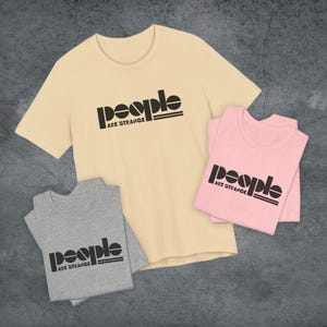 May include: Three t-shirts in cream, pink, and gray, each with the text "People Are Strange" in black. The shirts are folded or laid flat, showcasing the graphic design. The t-shirts are short-sleeved.
