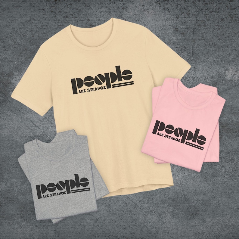May include: Three t-shirts in cream, pink, and gray, each with the text "People Are Strange" in black. The shirts are folded or laid flat, showcasing the graphic design. The t-shirts are short-sleeved.