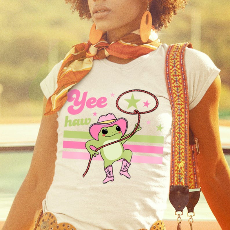 Pu&ograve; includere: Maglietta bianca con una rana verde che indossa un cappello da cowboy rosa e stivali. La rana tiene un lazo e il testo "Yee haw" &egrave; stampato sulla maglietta.