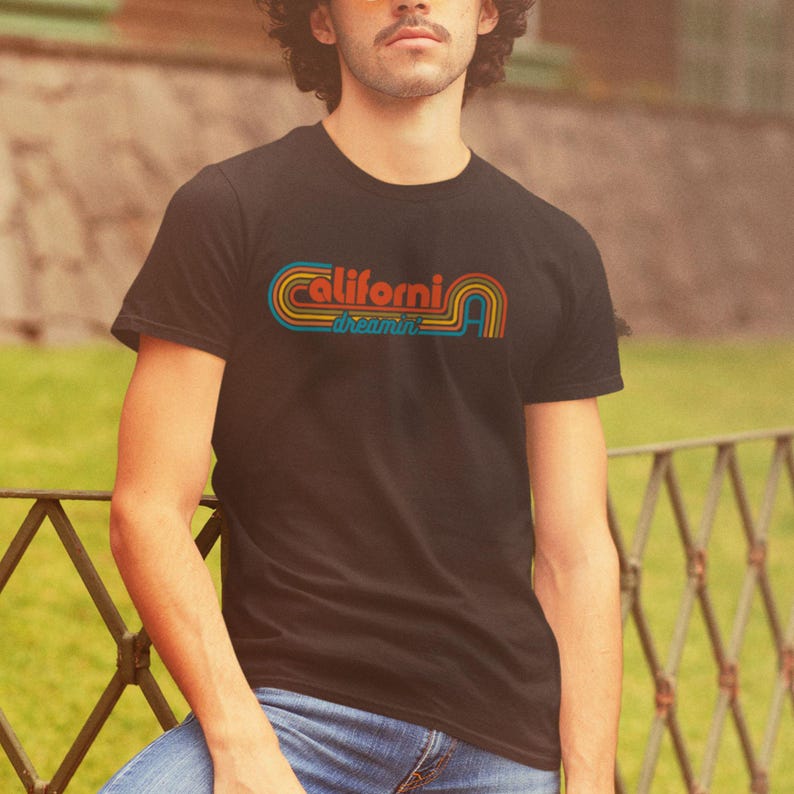 Op de afbeelding: Een zwart T-shirt met een retro-grafische afbeelding met de tekst "California Dreamin'" in regenboogkleuren.