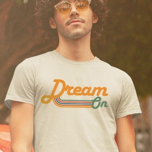K&ouml;nnte beinhalten: Eine Person tr&auml;gt ein hellbeiges T-Shirt mit dem Text "Dream On" in einer Retro-Schriftart mit bunten Streifen. Der Text ist in Orange, Gr&uuml;n, Blau und Rot.