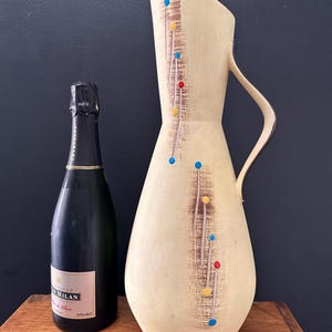 Puede incluir: Una jarra alta de cerámica color crema con un asa curva y un patrón decorativo de puntos y líneas de colores. Una botella de champán verde oscuro está al lado de la jarra. La jarra mide unos 30 cm de alto.