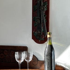 Pode incluir: Uma garrafa de vinho com um rótulo escuro e uma rolha está sobre uma superfície de madeira ao lado de duas taças de vinho gravadas. Um abridor de vinho decorativo montado na parede com detalhes de uvas e folhas é visível.
