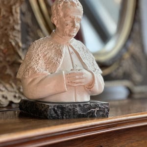 May include: A cream-colored bust sculpture of a person with a textured cape and clasped hands, mounted on a dark, rectangular base. The sculpture is set on a wooden surface, with a blurred mirror in the background.