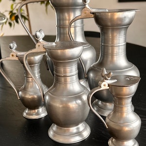 Könnte beinhalten: Eine Sammlung von Zinnkrügen in verschiedenen Größen. Jeder Krug hat einen Griff, einen Klappdeckel und eine dekorative Verzierung. Die Krüge sind auf einer dunklen Oberfläche angeordnet, die ihren metallischen Glanz und ihr klassisches Design hervorhebt.