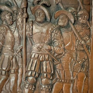 Può includere: Un'intagliatura in legno dettagliata raffigurante un gruppo di uomini in abiti storici. Le figure indossano abiti d'epoca, tra cui cappelli, armature e calzoni. Alcune figure impugnano armi, come spade e lance. L'intaglio è nei toni del marrone.