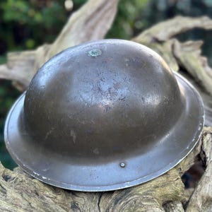 Puede incluir: Un casco de acero verde oliva vintage, con una corona redondeada y un ala ancha. El casco muestra signos de desgaste y edad, con arañazos y un acabado ligeramente deslustrado. Un pequeño emblema circular es visible.