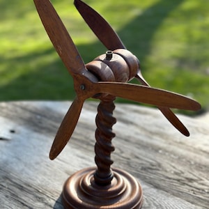 Op de afbeelding: Een houten propellermodel met drie bladen, gemonteerd op een gedraaide kolom en een cirkelvormige basis. De propellerbladen zijn warmbruin van kleur en de basis is donkerder bruin. Het model wordt op een houten oppervlak getoond.