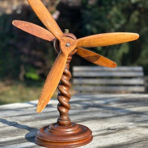 Peut inclure: Maquette d'hélice en bois à quatre pales, montée sur une colonne torsadée marron foncé et une base circulaire. Les pales sont d'une teinte marron plus claire, et le modèle est exposé sur une surface en bois.