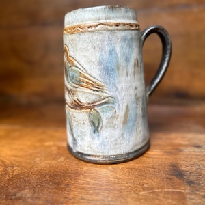 Puede incluir: Una taza cilíndrica de cerámica con un esmalte moteado en azul claro y blanco. El exterior presenta un relieve de un pájaro posado en una rama. Un asa de color marrón oscuro está unida al lateral y una banda decorativa rodea el borde.