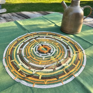 May include: A round, handmade crochet doily with a geometric design in shades of brown, yellow, and cream. The doily is placed on a green tablecloth, with a brown ceramic pitcher in the background.
