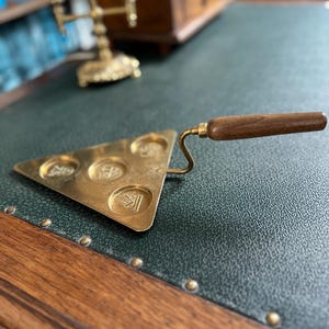 Könnte beinhalten: Ein Käsemesser aus Messing mit Holzgriff. Das Messer hat eine dreieckige Klinge mit vier kreisförmigen Vertiefungen. Der Griff ist dunkelbraun. Das Messer liegt auf einer grünen Oberfläche.