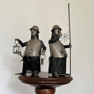 Op de afbeelding: Twee metalen sculpturen van middeleeuwse wachters. Eén houdt een lantaarn en een zwaard vast, de ander een speer met een spinnenwebontwerp. Beide figuren dragen helmen, harnassen en maliënkolders. De sculpturen zijn gemonteerd op een donkere houten voet.