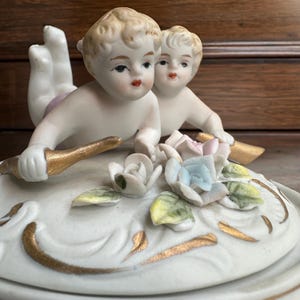 Pode incluir: Uma estatueta de porcelana branca com duas figuras de querubins com cabelo loiro e bochechas rosadas. Os querubins estão posicionados acima de um arranjo floral com detalhes dourados. As flores são brancas, azuis e rosas.