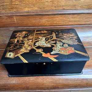 Peut inclure: Boîte rectangulaire noire avec un couvercle décoratif orné d'un motif d'inspiration asiatique doré et rouge. Le couvercle représente des personnages en vêtements traditionnels, sur un paysage avec des motifs géométriques. La boîte est posée sur une surface en bois.