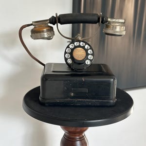 May include: An antique black telephone with a rotary dial and a black receiver. The phone sits on a small, round, black table with a wooden base. The phone has a vintage design.