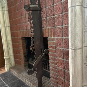 May include: A dark metal fireplace tool with a saw-like blade and decorative scrollwork. A chain hangs from the top of the tool. The tool is positioned in front of a red brick fireplace.