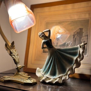 Peut inclure: Une figurine vintage en porcelaine d'une danseuse dans une robe fluide bleue et blanche avec des bordures dorées. Une lampe de bureau en laiton avec un abat-jour en verre orange et blanc illumine la scène. Une image encadrée est en arrière-plan.
