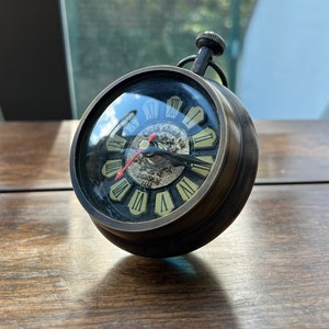 Könnte beinhalten: Eine kugelförmige Uhr im Vintage-Stil mit dunkler Bronzeoberfläche. Das Zifferblatt zeigt römische Ziffern und einen roten Sekundenzeiger. Sie hat einen kleinen, kreisförmigen Griff und steht auf einer Holzoberfläche. Der Durchmesser der Uhr beträgt ca. 8 cm.