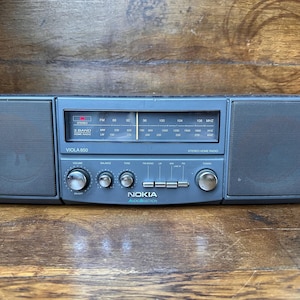 Può includere: Una radio stereo domestica Nokia Viola 850 grigio scuro con due altoparlanti integrati sui lati, un display di sintonizzazione analogico centrale con bande FM, MW e LW, manopole per volume e bilanciamento e quattro pulsanti per la selezione.