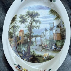 Może przedstawiać: Owalny ceramiczny półmisek z malowaną sceną krajobrazu zamku, rzeki i postaci w łodzi. Obrzeże jest kremowe z kwiatowymi i liściastymi akcentami w odcieniach czerwieni, żółci i fioletu.