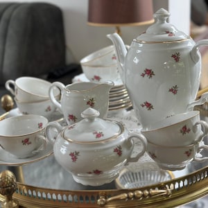Può includere: Un servizio da tè in porcellana vintage con teiera, zuccheriera, lattiera, tazze e piattini. Il set bianco è decorato con piccoli grappoli di rose rosse e finiture dorate. Il set è esposto su un vassoio di vetro con bordo dorato.
