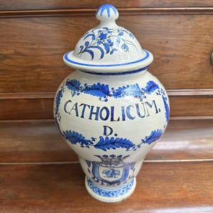 Può includere: Un vaso da farmacia in ceramica bianca con coperchio, decorato con motivi floreali blu e la parola "CATHOLICUM". Il vaso presenta uno stemma decorativo ed è probabilmente un pezzo vintage o antico. Il vaso è alto circa 30 cm.
