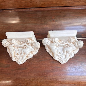 Könnte beinhalten: Zwei cremefarbene, dekorative Wandleuchten mit einem Cherub-Design. Jede Wandleuchte hat eine rechteckige Oberseite und eine geformte Basis mit drei Cherubim. Die Cherubim sind in einer spielerischen Pose dargestellt, mit detaillierten Gesichtszügen und fließenden Draperien.
