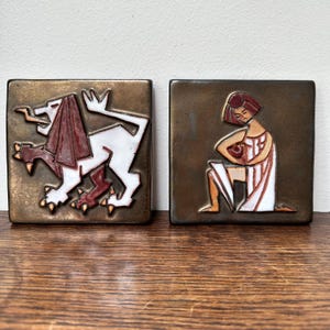 May include: Two square, bronze-coloured tiles with relief designs. One shows a white lion with a red mane and legs. The other depicts a person in a white robe holding a red object. The tiles are likely decorative.