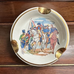 May include: A vintage, round ashtray with a cream-colored base and a gold rim. The ashtray features a colorful illustration of soldiers on horseback and a cannon. The ashtray has three gold-colored rests for cigarettes.