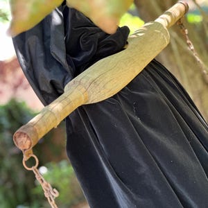 Op de afbeelding: Een lichtbruine houten deegroller, bevestigd aan een zwarte stof. De deegroller wordt opgehangen door een metalen ketting. De achtergrond is wazig met groene en bruine tinten.