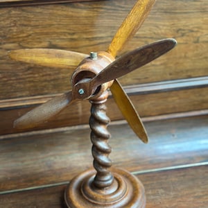 Könnte beinhalten: Ein Vintage-Modell eines Holzpropellers mit drei Blättern. Die Blätter sind hellbraun und an einer zentralen Nabe befestigt. Der Propeller sitzt auf einem gedrehten, geschnitzten Holzständer und einem kreisförmigen Sockel. Das Holz hat ein warmes, gealtertes Aussehen.