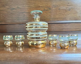 Op de afbeelding: Een vintage likeur karaf set met een gouden basis en horizontale witte strepen. De set bevat een karaf met een stop en zes bijpassende borrelglaasjes. De borrelglaasjes en de stop hebben een groene accentkleur.