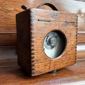 Könnte beinhalten: Eine Vintage-Holzkiste mit Ledergriff und rundem Fenster mit Zifferblatt. Die Kiste ist mit Schwalbenschwanzverbindungen konstruiert und hat eine dunkelbraune Oberfläche. Das Zifferblatt zeigt "OUTLET TIME".