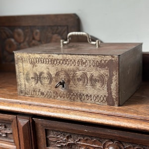 May include: A rectangular, antique metal box with a silver handle and keyhole. The box is brown with a decorative circular pattern and a key in the lock. The box is sitting on a wooden surface.