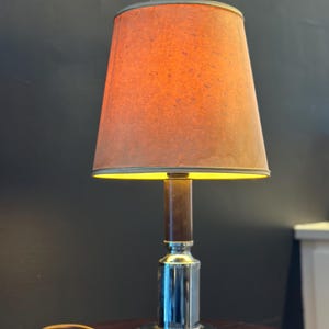 Può includere: Una lampada da tavolo vintage con un paralume arancione caldo. La lampada ha una base cromata e una colonna in legno marrone. La lampada è su un tavolo di legno scuro. La lampada è collegata e pronta all'uso.