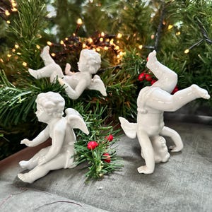 Puede incluir: Tres figuras de querubines de porcelana blanca en varias poses, con pequeñas alas y pelo rizado. Los querubines están colocados sobre un fondo de follaje verde y luces parpadeantes, con bayas rojas.