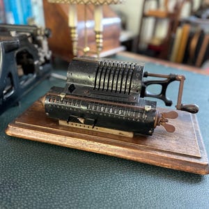 Op de afbeelding: Een antieke zwarte mechanische rekenmachine met meerdere nummerplaten en een slinger, gemonteerd op een houten voet. De rekenmachine toont het nummer 00256506000. Een bureaulamp en een typemachine staan op de achtergrond.