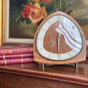 Op de afbeelding: Een vintage houten klok met een driehoekige vorm en een lichtgekleurde wijzerplaat. De klok heeft goudkleurige wijzers en een glazen afdekking. Hij staat op kleine metalen pootjes. Op de achtergrond een schilderij van rozen en een stapel rode boeken.