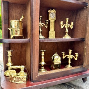 Può includere: Un mobiletto in legno in miniatura con più ripiani che espongono oggetti decorativi color oro. Gli oggetti includono un orologio, candelabri, una stufa e un ferro da stiro. Il mobile ha una finitura marrone scuro ed è ambientato su uno sfondo verde.